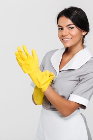 Close-up photo of young smiling brunette maid in uniform putting on yellow rubber gloves while standing and looking at camera, isolated on white background